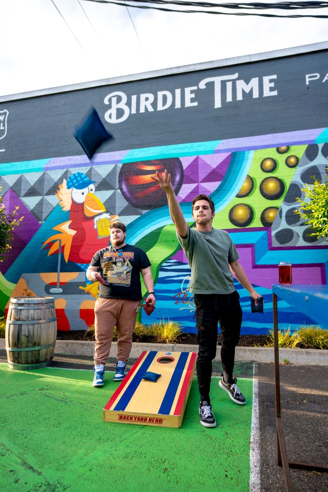 Two men playing cornhole in front of a colorful mural wall at Birdie Time mini golf. 