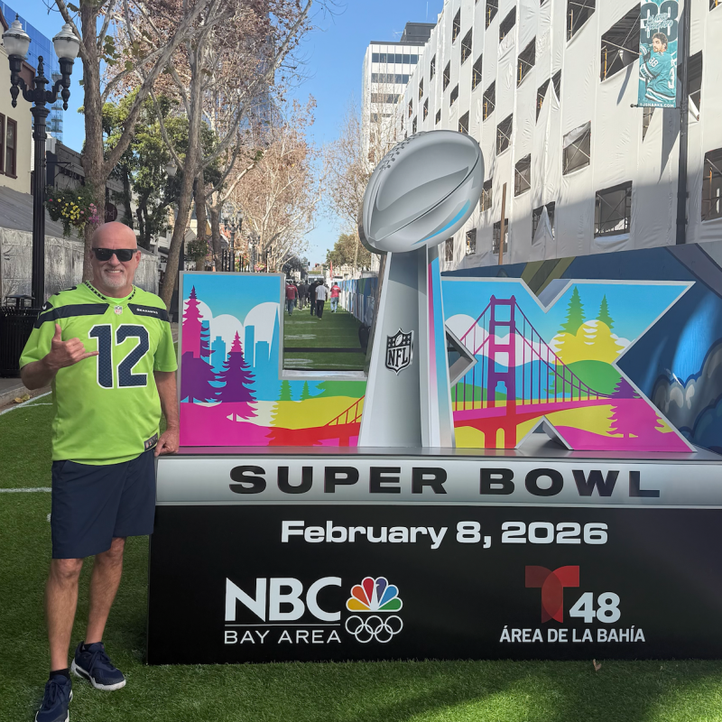 Man in sports jersey stands by a Super Bowl display in an outdoor urban setting.