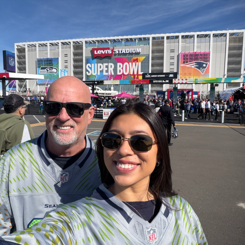 Two people smiling in front of Levi's Stadium with Super Bowl signage.