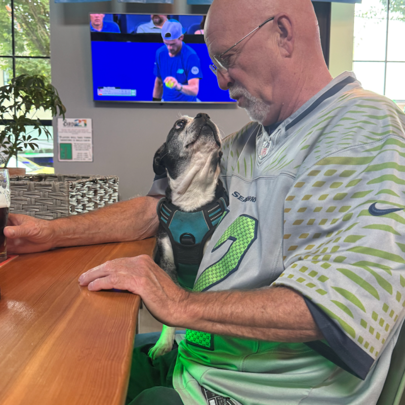 Man in sports jersey holds small dog at a bar table.